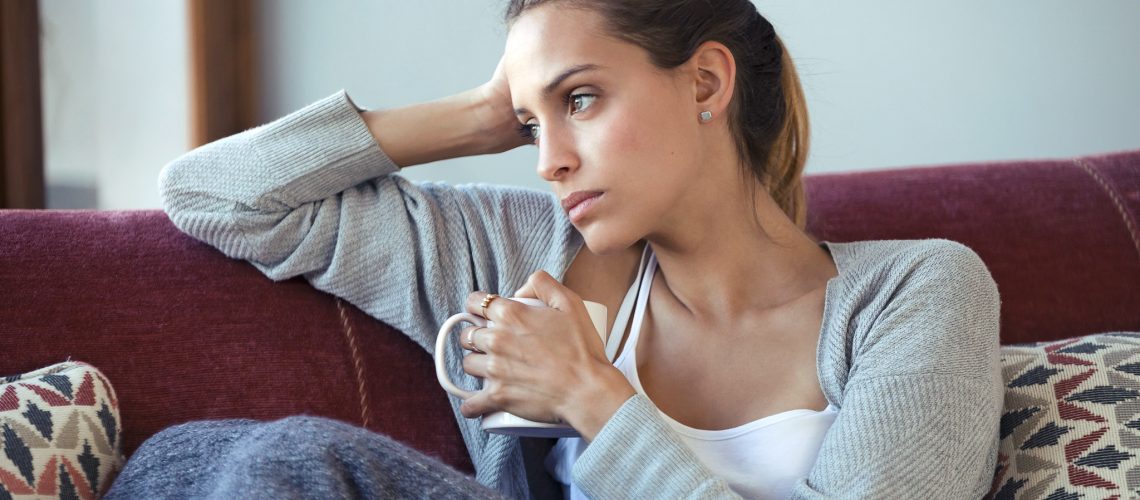 Shot of depressed young woman thinking about her problems while drinking coffee on sofa at home.